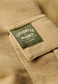 Tela color beige con textura visible; una etiqueta verde que dice "Superdry Co. Machined" con texto blanco, cosida en un bolsillo.