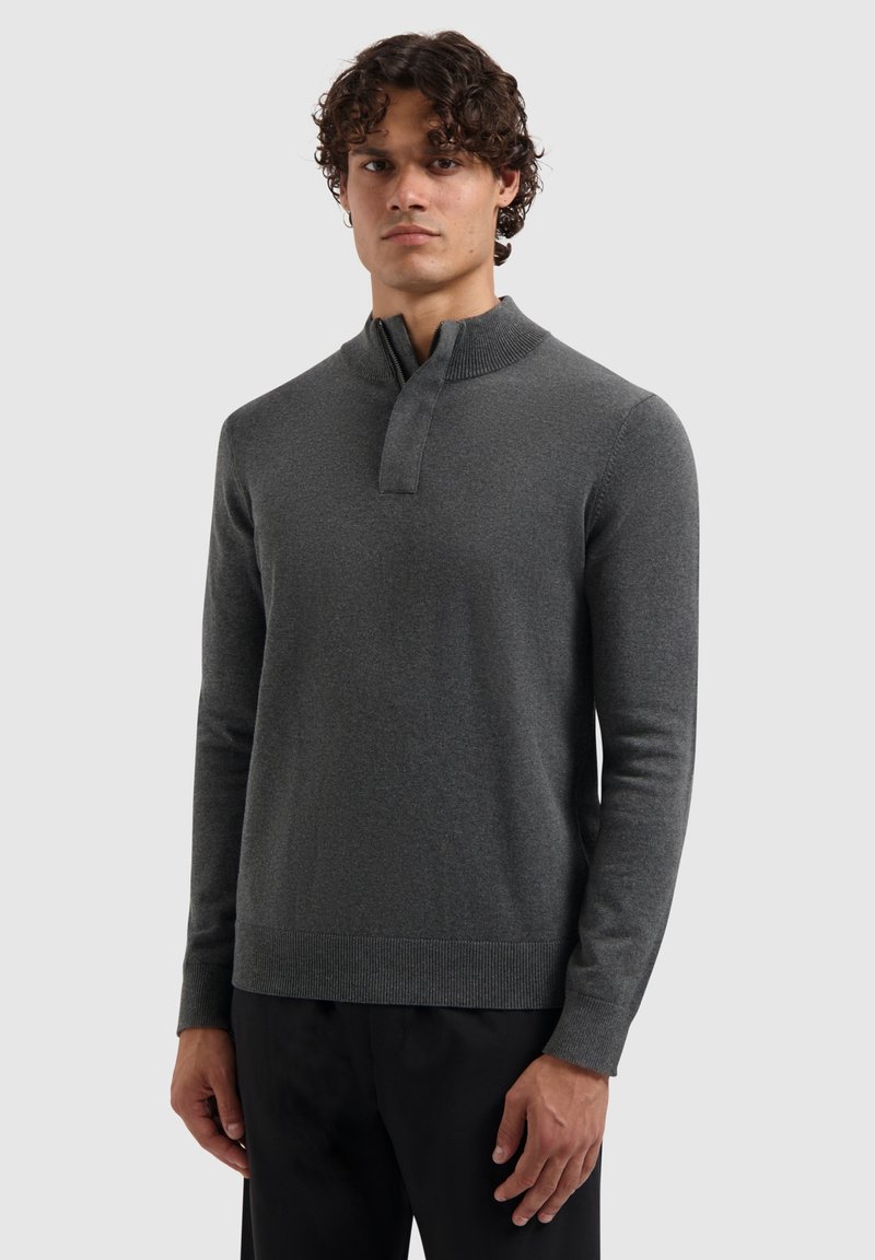 Jonge man draagt een donkergrijze, lange mouwen pullover met een geribde opstaande kraag en een gedeeltelijke ritssluiting aan de voorkant, gecombineerd met zwarte broek.