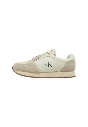 Zapatillas deportivas con un upper sintético color crema y acentos de ante beige, que presentan una suela de goma, cierre con cordones y un logotipo prominente en el lateral.