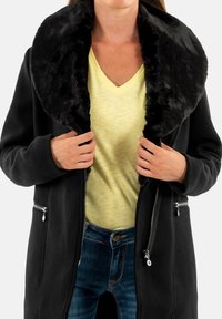Manteau noir avec un col en fausse fourrure ; dispose de deux poches zippées et d'une coupe décontractée. Porté sur un haut à col en V jaune clair et un jean en denim.