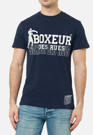 Uomo che indossa una t-shirt blu navy con testo bianco "BOXEUR DES RUES" e silhouette di pugilato, abbinata a jeans azzurri chiari.