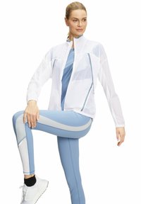 Witte lichte jas met een rits aan de voorkant, gecombineerd met lichtblauwe leggings met grijze accenten en een mesh-paneel. Atletische schoenen zichtbaar.
