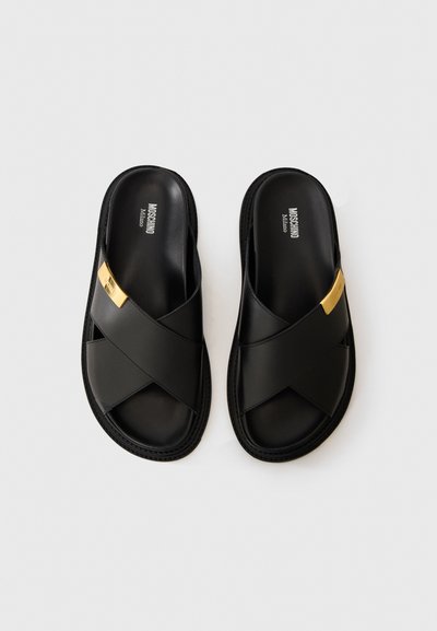 Svarta lädertofflor från Moschino med korskorsade remmar och detaljer i guld på vit bakgrund.