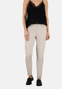 Pantaloni beige slim-fit con una texture liscia, dotati di una chiusura lampo frontale, passanti per la cintura e tasche laterali, abbinati a un top nero con orlo in pizzo.