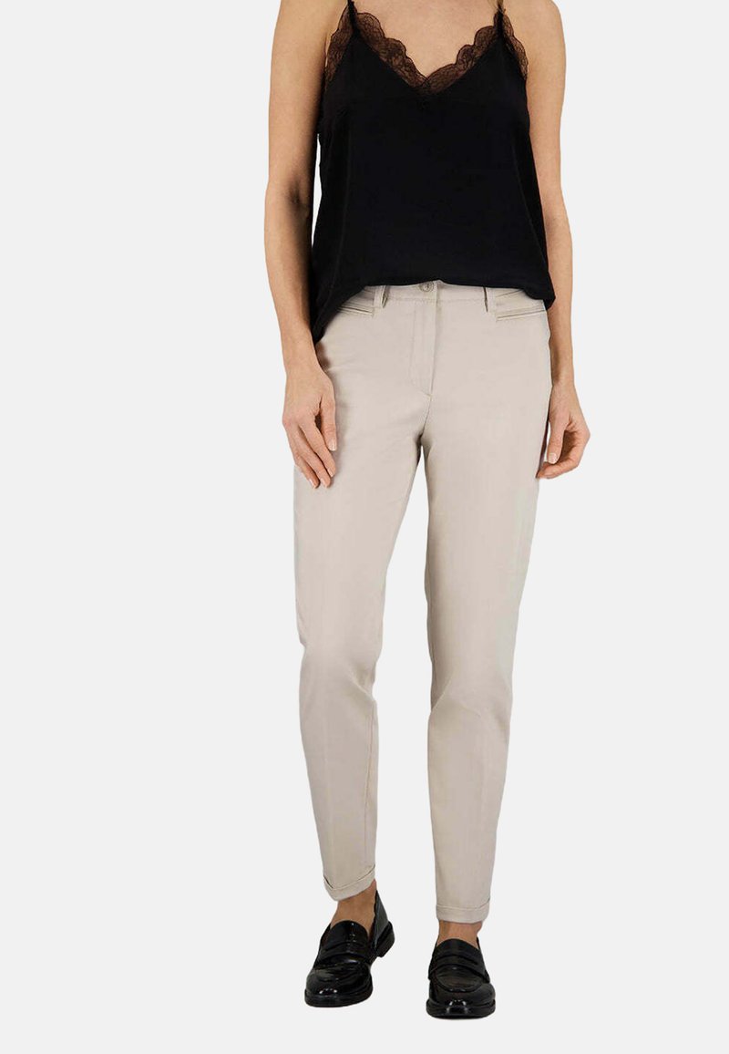 Pantaloni beige slim-fit con una texture liscia, dotati di una chiusura lampo frontale, passanti per la cintura e tasche laterali, abbinati a un top nero con orlo in pizzo.