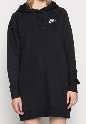 Personne portant un long sweat à capuche noir Nike avec une poche avant et un logo blanc sur la poitrine, debout devant un fond uni.