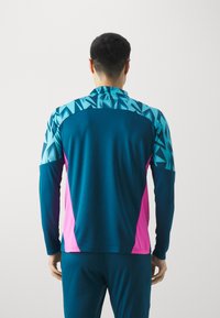 Langärmliges Sportshirt mit einem türkisfarbenen geometrischen Muster auf der Oberseite und leuchtend pinken Akzenten, aus leichtem Stoff gefertigt.