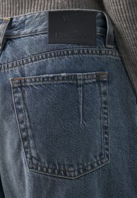 Achteraanzicht van een blauwe spijkerbroekzak met oranje stiksels en een zwart leren merklapje met de tekst "Won Hundred."