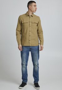 Chemise beige à manches longues avec un col, dotée de deux poches poitrine, associée à un jean bleu décoloré et des baskets noires.