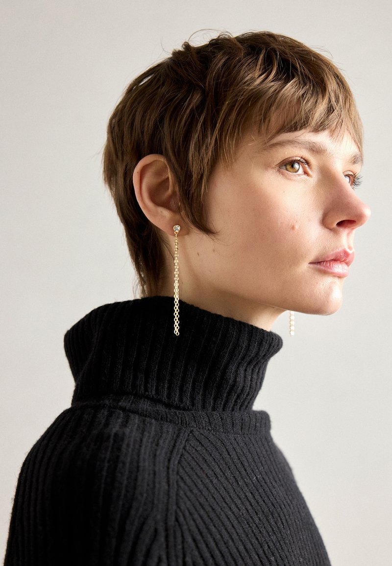 Gullige dråpeøredobber med kjededesign, som har en liten stud og lange hengende elementer. Modellen har på seg en svart ribbet turtleneck-genser.