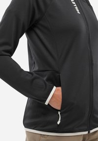Schwarze Outdoor-Jacke aus strukturiertem Stoff, mit durchgehendem Reißverschluss, Seitentaschen und weißen Saumabschlüssen an den Bündchen. Logo auf der linken Brust.