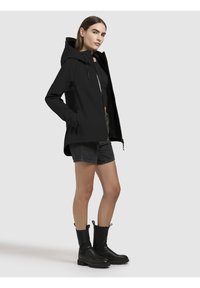 Zwarte waterdichte jas met capuchon, frontale rits en verborgen pockets, gecombineerd met grijze shorts en zwarte dikke enkellaarsjes.
