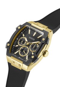 Montre Guess noire et dorée avec un boîtier rectangulaire, un bracelet en caoutchouc noir texturé et plusieurs cadrans pour la date et l'heure.