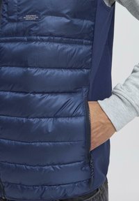 Muškarac nosi mornarsko plavi rameni prsluk sa rukom u zatvorenoj bočnoj džepu, a na prsima je vidljiva oznaka s tekstom brenda.