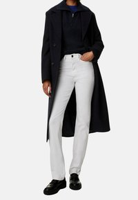 Marine overcoat, gestructureerde donkere trui met ritssluiting, witte straight-leg jeans en zwarte loafers. Voorzien van knopen en zijzakken.