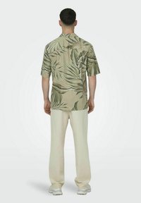 Korte mouwen shirt in beige met een groen palmblad patroon; gecombineerd met lichtgekleurde, losse broek en neutrale sportschoenen.