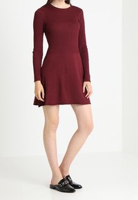 Abito bordeaux lungo fino al ginocchio con maniche lunghe e texture a coste nella parte superiore, e gonna svasata. Indossato con mocassini neri in vernice con borchie.