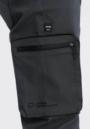 Pantalons cargo synthétiques gris avec une poche zippée, des détails cousus et une étiquette noire avec le texte "STAY REAL".