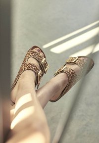 Sandále s leopardím vzorom, so zdravotnými zlatými prackami, otvorenou špičkou a plochou podrážkou. Materiál pôsobí štruktúrovane, s svetlohnedými a tmavohnedými akcentmi.