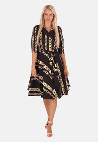 Frau mit langen blonden Haaren, die ein schwarzes knielanges Kleid mit goldenen Kettenmustern und hautfarbene High Heels trägt, steht vor einem weißen Hintergrund.