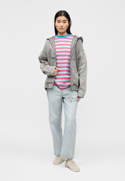 Νέα γυναίκα στέκεται, φορώντας ανοιχτό γκρι μπουφάν με κουκούλα, ροζ και μπλε ριγέ πουκάμισο, ανοιχτού μπλε τζιν και μπεζ αθλητικά παπούτσια σε λευκό φόντο.