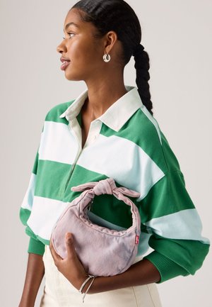 Frau mit geflochtenen Haaren, die ein grün-weiß gestreiftes Hemd trägt und eine kleine hellviolette Stoffhandtasche mit einer gebundenen Schleife am Griff hält.