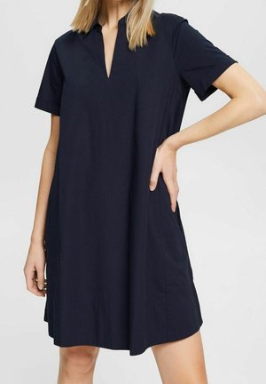 Frau trägt ein locker sitzendes, knielanges schwarzes Kleid mit kurzen Ärmeln und V-Ausschnitt, posiert mit einer Hand in der Hüfte.