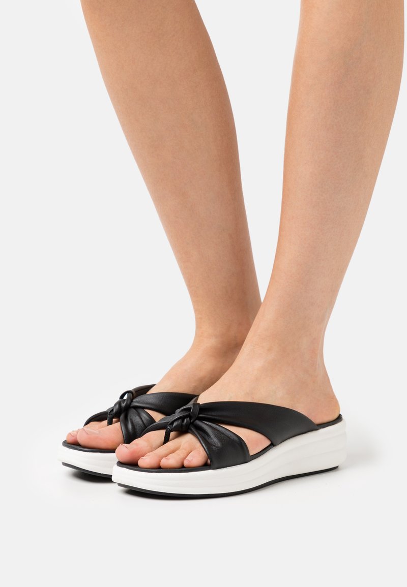 Pies que llevan sandalias de tiras anudadas negras con suelas de plataforma blancas sobre un fondo blanco.