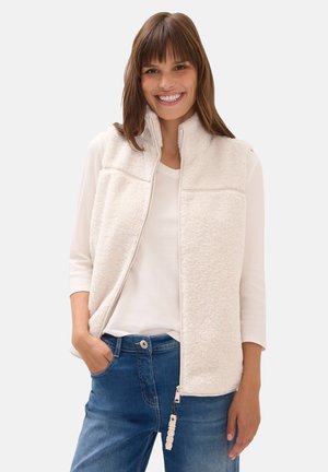 Hengittävä fleece-liivi vaaleassa kermassa, jossa on korkea kaulus, vetoketjusuljenta ja kaksi etutaskua. Yhdistettynä valkoiseen paitaan ja sinisiin farkkuihin.