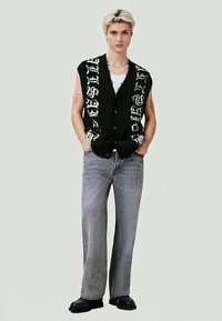 Jovem com cabelo curto loiro, vestindo um colete preto sem mangas com botões e letras brancas, calças cinzas largas e sapatos pretos, de pé com as mãos nos bolsos.