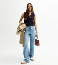 Marinblå stickad väst med knappar, ljusblå vida jeans, beige trenchcoat hängande över armen, brun väska och vita sneakers med bruna accenter.