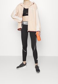 Světle růžová zipová mikina s kapucí a bílými pruhy, černá sportovní podprsenka, černé legíny se šedými akcenty a černé tenisky, držící oranžovou láhev na vodu.