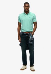 Polo shirt vert menthe avec un col, jeans sombres tissés, et flanelle à carreaux nouée à la taille. Baskets beiges avec des accents bruns complètent le look.