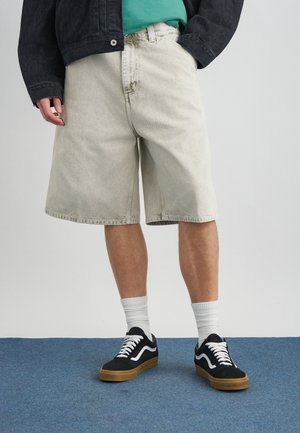 Persona vistiendo pantalones cortos de mezclilla beige claro de gran tamaño, zapatillas negras con cordones blancos, calcetines blancos hasta la pantorrilla, camiseta verde azulado y chaqueta oscura.