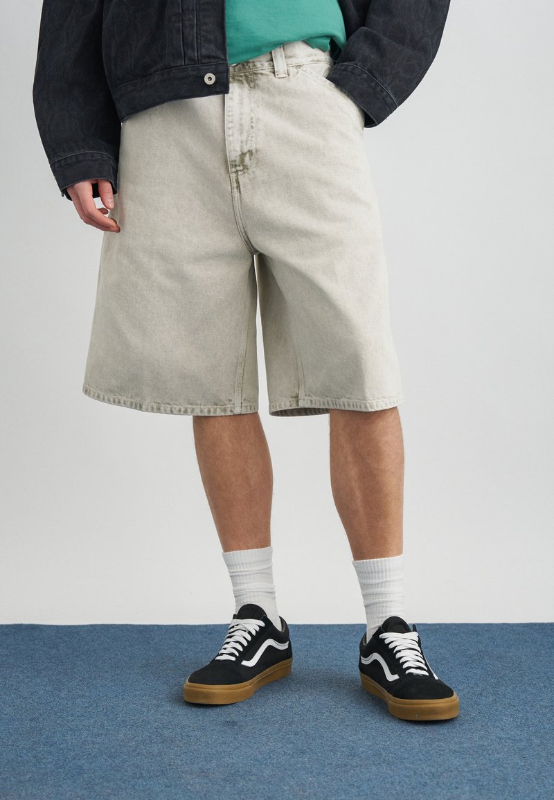 Άτομο που φοράει ανοιχτό μπεζ φαρδιά τζιν σορτς, μαύρα αθλητικά παπούτσια με λευκά κορδόνια, λευκές κάλτσες μέχρι τον αστράγαλο, τιρκουάζ πουκάμισο και σκούρο σακάκι.