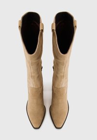 Bottes en suede beige montantes jusqu'aux genoux avec bout pointu, texture lisse et anses de traction. Présente des accents brodés sur la tige pour plus de détails.