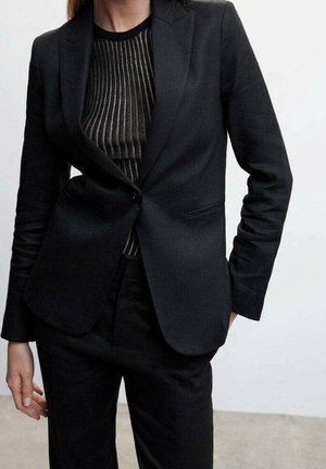 Femme portant un blazer noir ajusté sur un top rayé vertical noir et marron, associé à un pantalon noir taille haute.