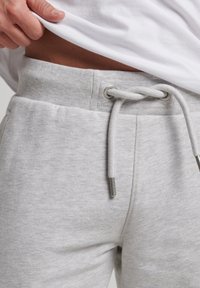 Acercamiento de una persona levantando una camiseta blanca, revelando la cinturilla y el cordón de unos pantalones de chándal gris claro.