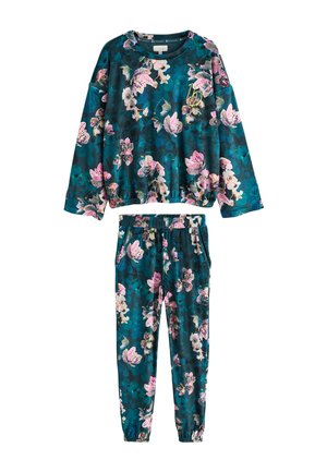 Kvinde i mørkt blomsterprintet nattøj står barfodet ved en gardin, holder en hvid kop og kigger eftertænksomt til siden.