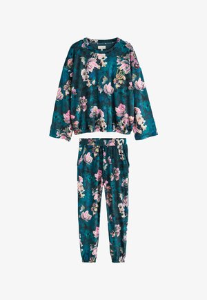Vrouw in donkere bloemrijke loungewear staat blootsvoets bij een gordijn, houdt een witte mok vast en kijkt nadenkend naar de zijkant.