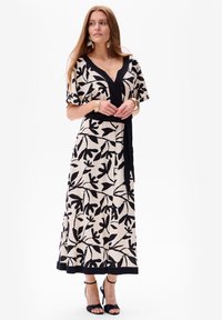 Maxi-jurk in roomkleur met zwart bladmotief, V-halslijn, korte mouwen, cinched taille en zwarte accent bij de zoom. G gedragen met zwarte sandalen met bandjes.