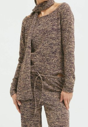 Mujer con un suéter de punto jaspeado en tonos morado y beige, pantalones a juego y una bufanda larga de punto colgada alrededor del cuello.