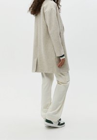 Manteau en laine beige clair à coupe décontractée, avec deux poches latérales. Associé à un pantalon crème et des baskets à accents vert foncé.