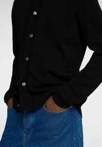 Svart strikket cardigan med sølvknapper, brukt over blå denimjeans. Viser en avslappet passform og teksturert stoff.