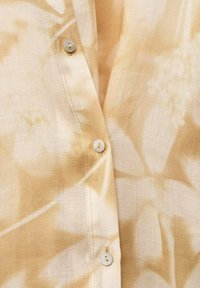 Camicia beige chiaro con motivo floreale; realizzata in tessuto morbido; presenta bottoni perlati e cuciture visibili lungo il pannello frontale.