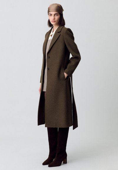 Cappotto di lana marrone con un design su misura, rever a contrasto, due bottoni, tasche laterali, indossato sopra un vestito beige e abbinato a stivali scuri fino al ginocchio.