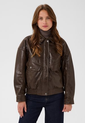 Femme aux longs cheveux bruns portant une veste en cuir marron foncé, un col roulé gris et un jean bleu foncé, debout devant un fond uni.