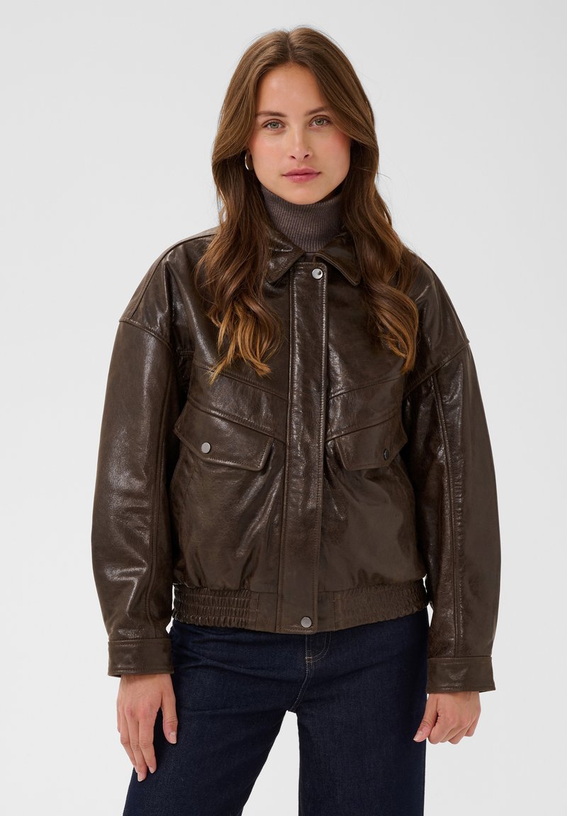 Femme aux longs cheveux bruns portant une veste en cuir marron foncé, un col roulé gris et un jean bleu foncé, debout devant un fond uni.
