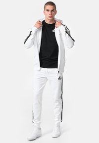 Vit träningsoverall bestående av en zip-up hoodie och tapered byxor, svarta accenter på ärmar och byxor, i kombination med vita sneakers.