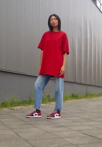 Camiseta roja de gran tamaño con un logo, combinada con jeans de pierna recta de color azul claro y zapatillas rojas, negras y blancas. Fondo gris.